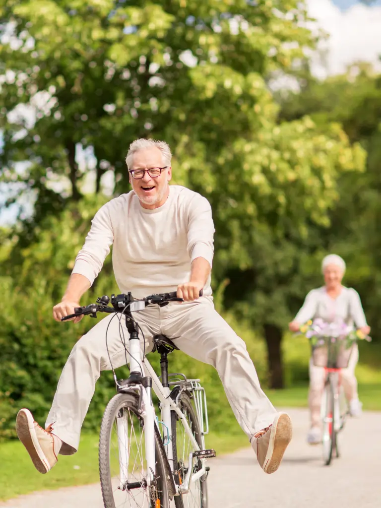 Ein Mann sitzt fröhlich auf seinem Fahrrad und hält die Füße hoch. Eine Frau fährt hinter ihm Fahrrad durch einen Park.
