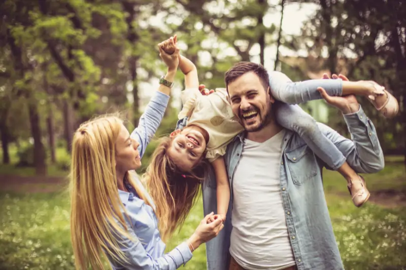 Eine Familien im Wald. Die Tochter liegt lachen über den Schultern des Vaters, die Mutter hält sie an den Händen.