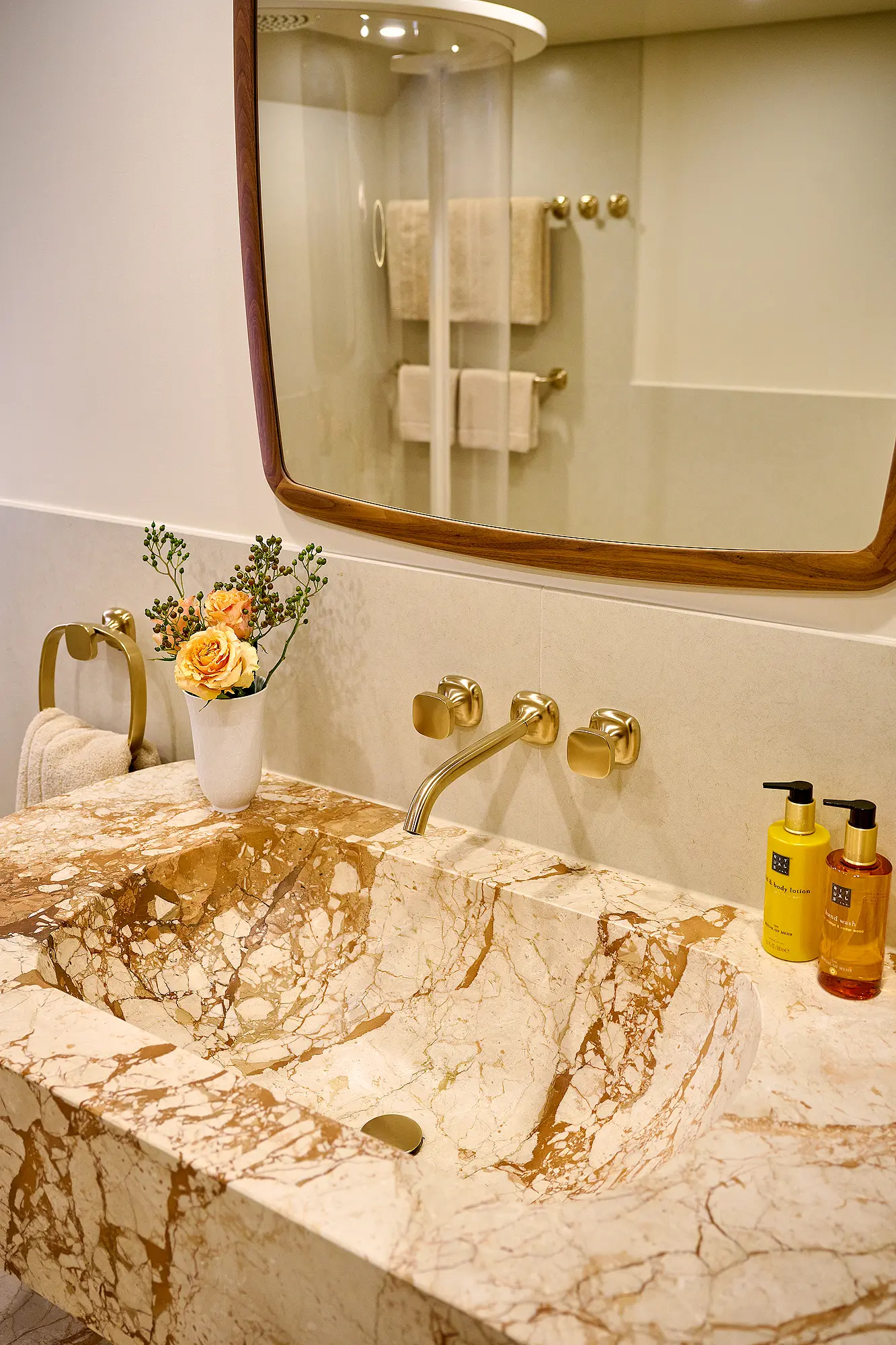 Badezimmer mit Waschtisch aus hellem, gemasertem Naturstein, goldfarbenen Armaturen und großem Spiegel.